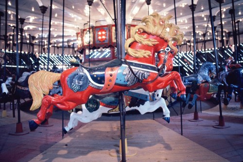 A vibrant red carousel horse with a flowing mane, set against a colorful, illuminated carousel backdrop.