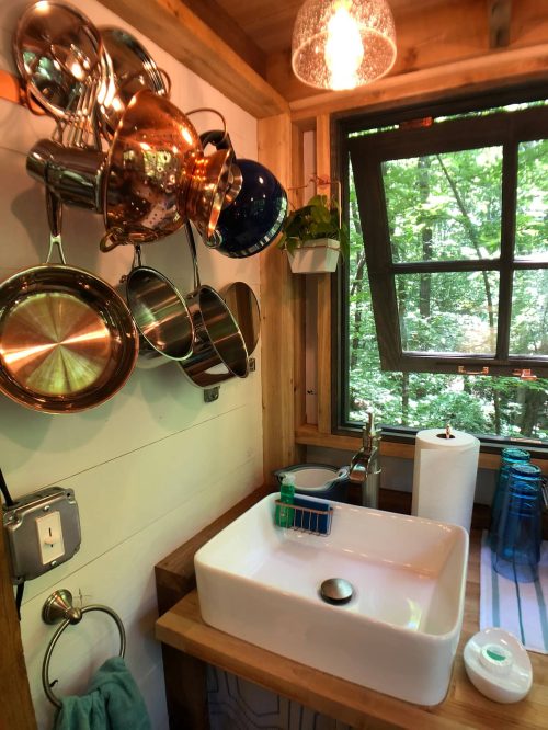 A cozy kitchen sink area with hanging pots, a window view of greenery, and wooden accents.