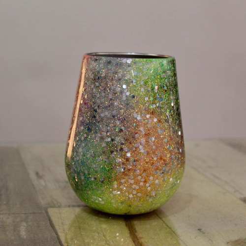 A shimmering, multicolored glitter cup with a gradient of green, gold, and orange tones on a wooden surface.