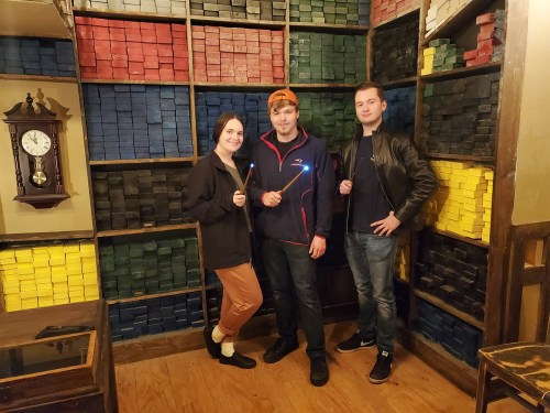 Three people stand in front of a colorful wall of stacked blocks, each holding a glowing wand.