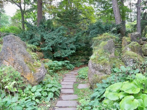 A serene garden path winding through lush greenery and moss-covered rocks, surrounded by trees.