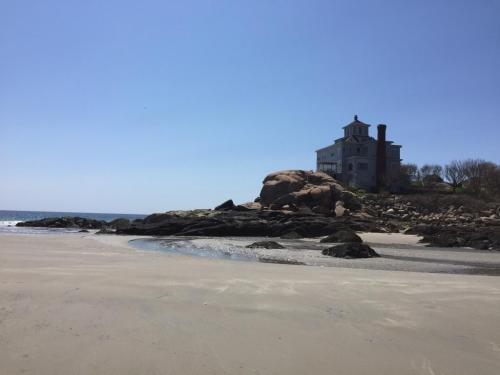 A sandy beach with a rocky shoreline and a large house perched on a hill under a clear blue sky.