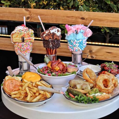 A table filled with colorful milkshakes, burgers, fries, salad, and onion rings, surrounded by greenery.