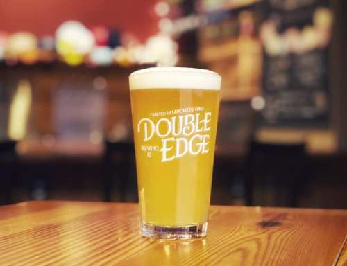 A glass of pale beer with a frothy head sits on a wooden table, featuring the logo of Double Edge Brewing.