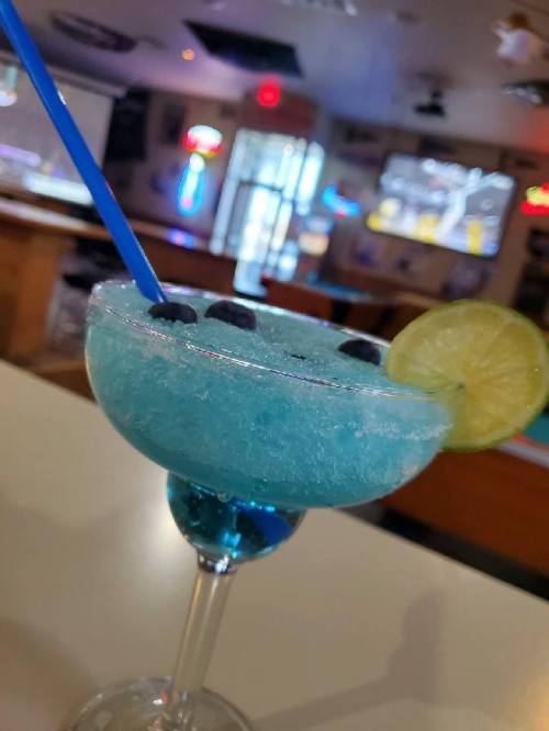 A blue frozen cocktail garnished with blueberries and a lime slice, served in a glass with a straw.
