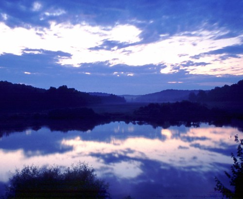 A serene river at dusk, reflecting a colorful sky with clouds and silhouettes of trees along the banks.