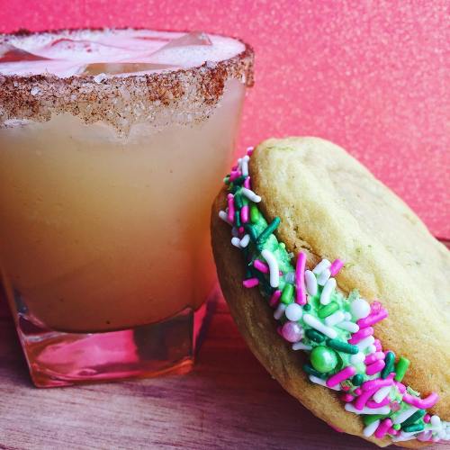 A colorful cookie with sprinkles next to a cocktail in a glass with a sugared rim, set against a glittery background.