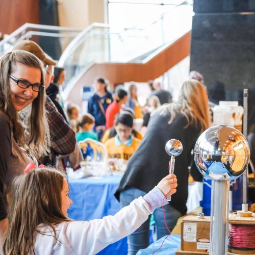 A child interacts with a science exhibit while adults and other children engage in activities in the background.