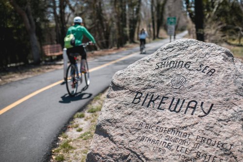 A stone marker for the "Shining Sea Bikeway" with cyclists riding on a paved path in the background.