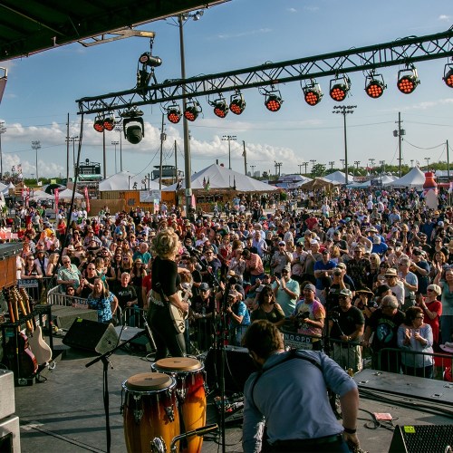 A lively outdoor concert scene with a large crowd enjoying a performance on stage under a clear blue sky.