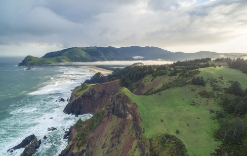Aerial view of a lush green coastline with cliffs, waves crashing, and misty mountains in the background.
