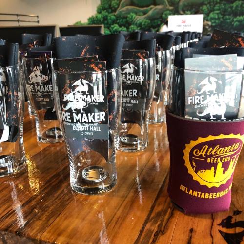 A table with glasses and koozies from Fire Maker Brewing Company and Atlanta Beer Bus, set in a brewery.