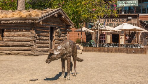 A bronze wolf sculpture stands in front of a rustic wooden cabin and a restaurant with outdoor seating.