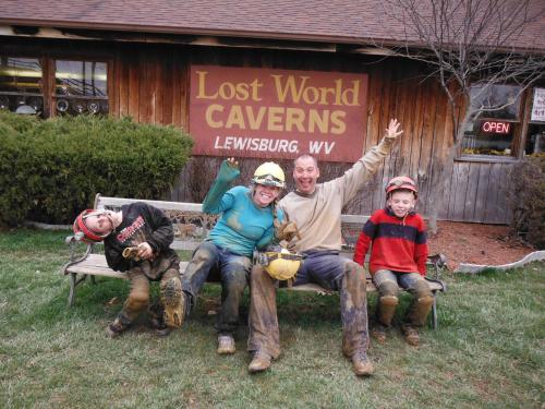 A group of four people, covered in dirt, joyfully pose on a bench in front of the Lost World Caverns sign in Lewisburg, WV.