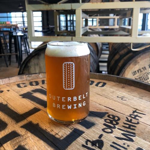 A glass of beer with the logo "Outerbelt Brewing" sits on a wooden barrel in a brewery setting.