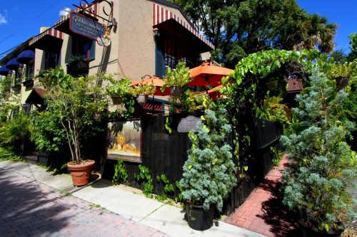 A charming outdoor dining area with greenery, potted plants, and orange umbrellas beside a quaint building.