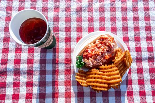A plate of pulled pork on a bun with coleslaw, served with waffle fries, next to a cup of iced drink on a checkered tablecloth.