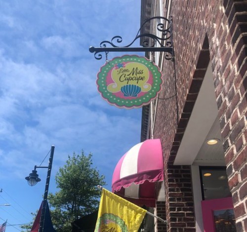 Sign for "Little Miss Cupcake" hanging from a brick building, with a blue sky and colorful flags in the foreground.