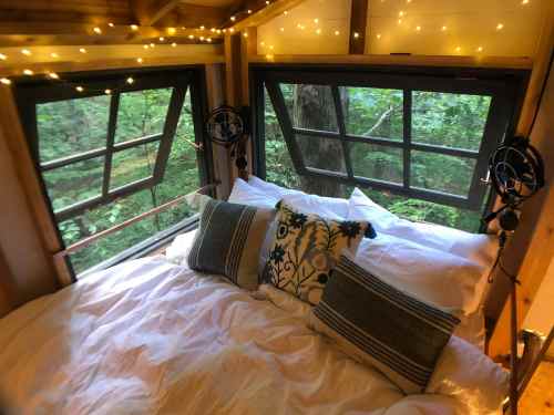 Cozy bedroom with a bed, decorative pillows, and large windows overlooking a lush green forest, illuminated by fairy lights.
