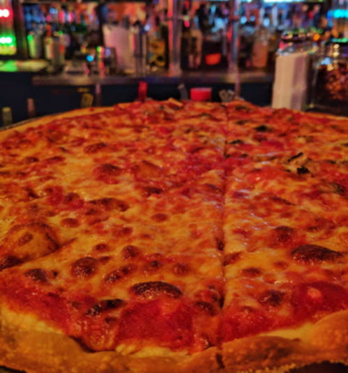 A close-up of a large cheese pizza with a golden crust, set against a blurred bar background.
