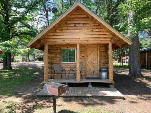 A small log cabin with a porch, two chairs, and a grill, surrounded by trees in a wooded area.