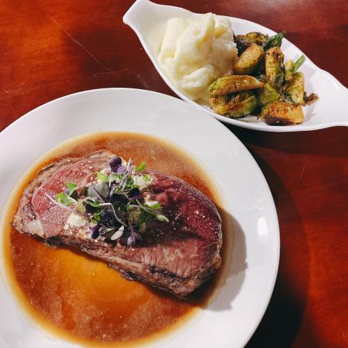 A plate of sliced roast beef with gravy, garnished with herbs, served with mashed potatoes and Brussels sprouts on the side.