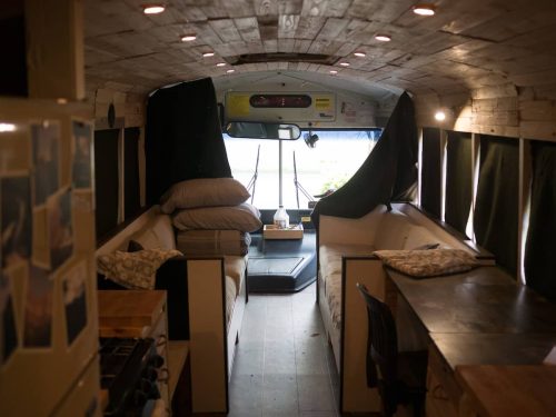 Interior of a converted bus, featuring cozy seating, wooden accents, and large windows with natural light.