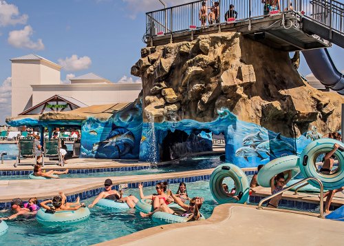 A vibrant pool scene with people relaxing on floats, a rock formation, and a water slide in the background.