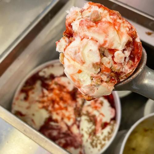 A scoop of ice cream with swirls of red sauce and chunks, held above a container of ice cream in a shop.