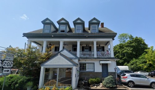 A large, historic house with a porch and multiple gabled roofs, surrounded by greenery and a small parking area.