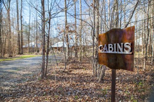 A rusted sign reading "CABINS" stands near a gravel path in a wooded area with bare trees and fallen leaves.