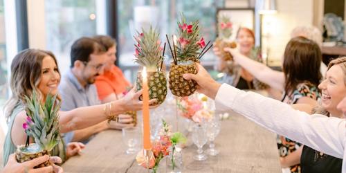 A group of people toasting with pineapple drinks at a festive dinner table adorned with flowers.