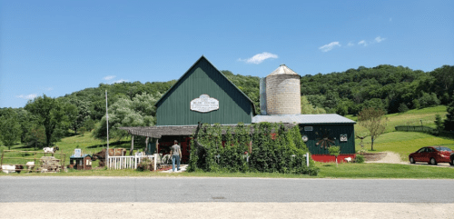 A green barn with a silo, surrounded by trees and a gravel road, with people outside on a sunny day.