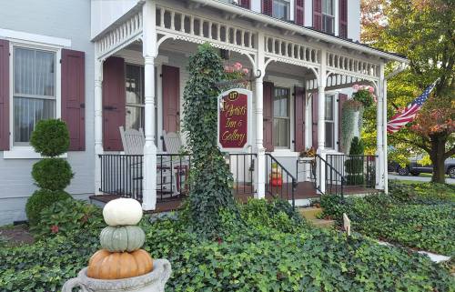 A charming inn with a porch, surrounded by greenery and pumpkins, featuring a sign that reads "Artist Inn & Gallery."