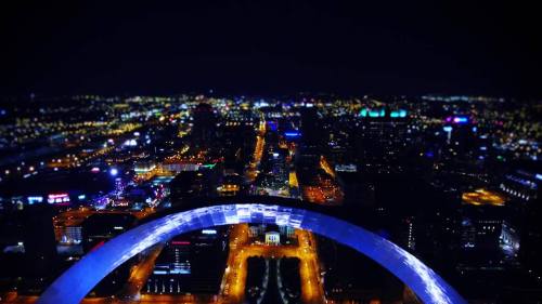 Aerial view of a city skyline at night, featuring illuminated buildings and streets beneath a large arch structure.