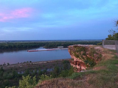 A scenic view of a river at sunset, with a rocky cliff in the foreground and a barge on the water.