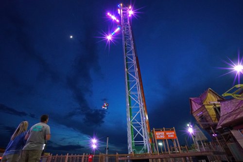 A tall amusement park ride illuminated at night, with two people watching from below and a crescent moon in the sky.