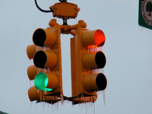 A traffic light with red and green signals, covered in icicles, against a gray sky.