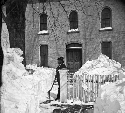 A person shovels snow in front of a brick building, with large snowdrifts and a white picket fence nearby.