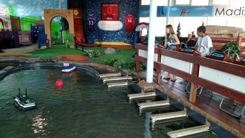 Children play with remote-controlled boats in a colorful indoor play area featuring a small water pond.
