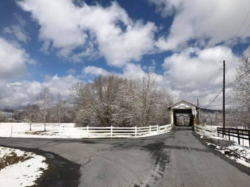 A snowy landscape featuring a covered bridge, trees, and a clear blue sky with fluffy clouds.