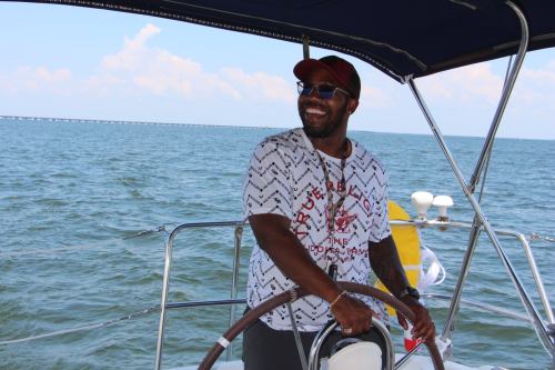 A smiling person steering a boat on a sunny day, with water and a bridge visible in the background.