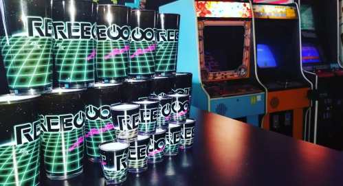 A stack of retro-themed cups with neon designs, next to vintage arcade machines in a dimly lit arcade.