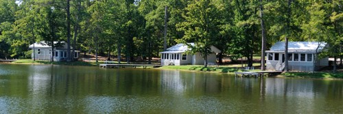 Three cabins by a calm lake, surrounded by lush green trees and reflecting in the water.