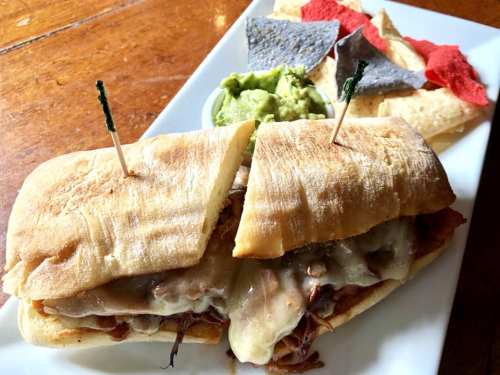 A toasted sandwich filled with melted cheese and meat, served with guacamole and colorful tortilla chips on the side.