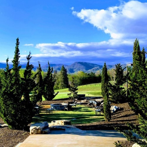 A scenic outdoor area with tables and chairs surrounded by trees, overlooking mountains under a blue sky.