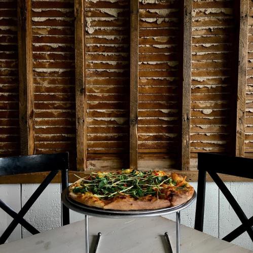 A pizza topped with greens sits on a stand in front of a rustic wooden wall.