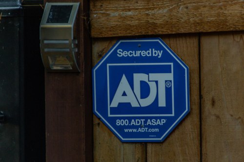 Blue security sign on wooden fence, stating "Secured by ADT" with contact number 800.ADT.ASAP.