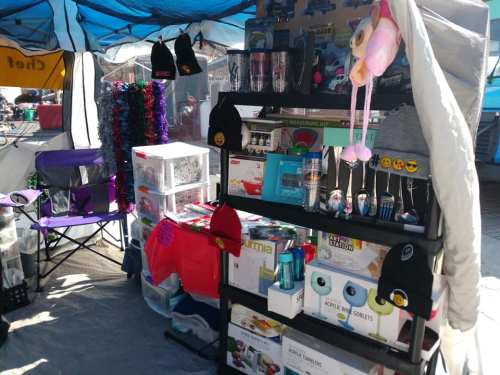 A market stall displaying various merchandise, including hats, kitchen items, and colorful decorations under a blue canopy.