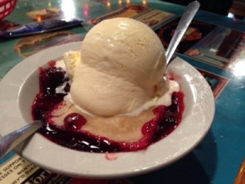 A scoop of vanilla ice cream on a bed of berry sauce, served in a small dish with two spoons.